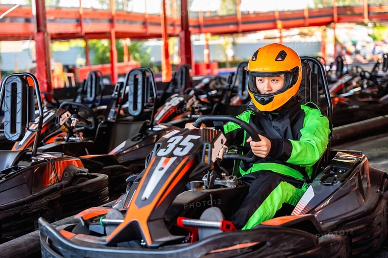 Young person wearing a bright green racing suit and orange helmet, sitting inside a go kart on the track, preparing for a race at an indoor karting facility