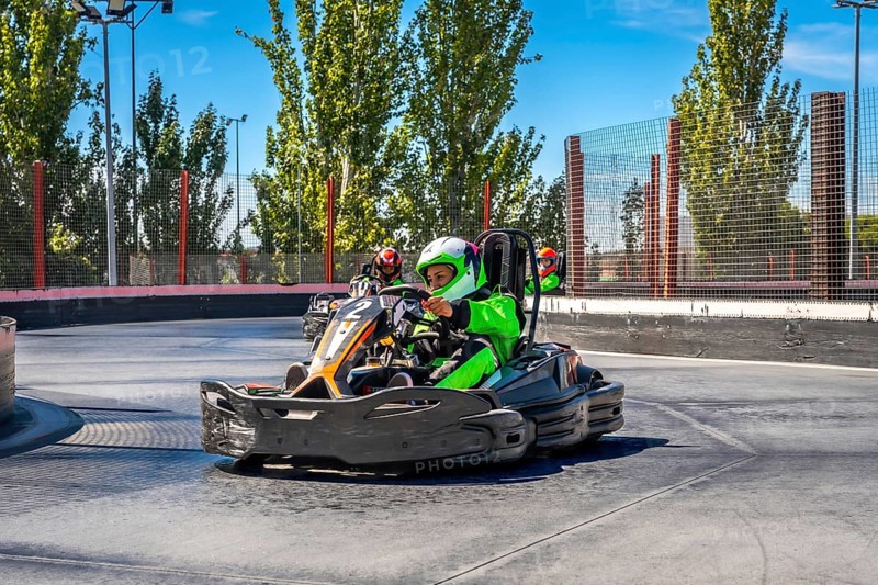 Drivers are speeding go karts around a curve on an outdoor asphalt track, wearing protective helmets and bright racing suits, enjoying the thrill of competition and motor sports on a sunny day