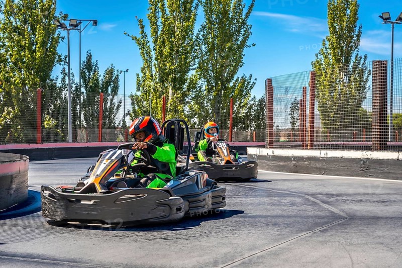Two children in helmets and protective gear race go karts on an outdoor track, enjoying the speed, competition and thrill of karting on a bright day