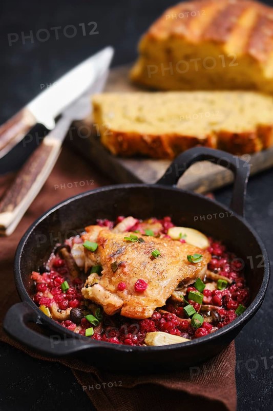 Roasted chicken thigh in cast iron skillet with lingonberry sauce. Slices of fresh bread are placed beside the pan on a wooden board. A knife and fork are shown