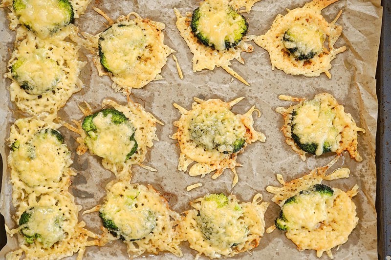 Broccoli pieces are smashed and spread on a baking sheet. They are topped with cheese and placed in an oven