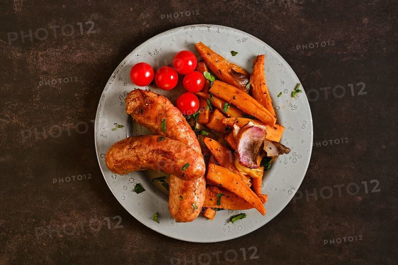 Top view of natural casing sausages on a plate alongside roasted sweet potatoes, onions, and cherry tomatoes. The meal shows a mix of colors with simple sides on a dark background
