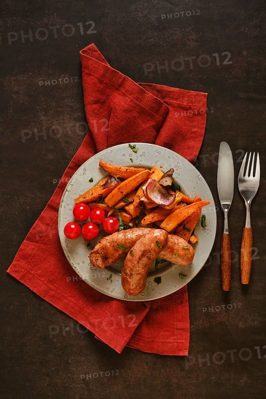 Natural casing sausages are placed on a green plate alongside roasted sweet potatoes and cherry tomatoes. A red napkin and utensils are arranged on a dark wooden table under soft light