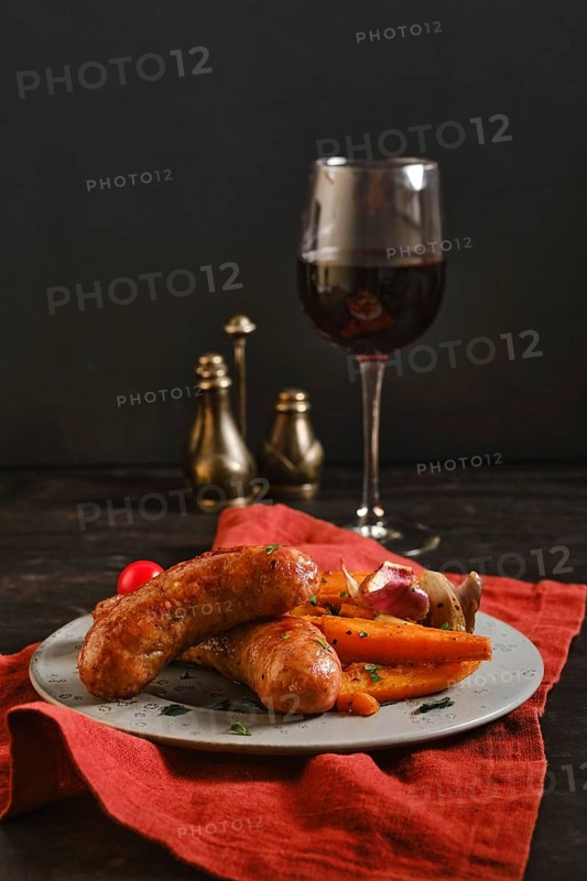 Sausages made with natural casings are placed on a plate with colorful vegetables. A glass of red wine stands nearby, creating a simple dining scene. The setting is warm and inviting for a meal