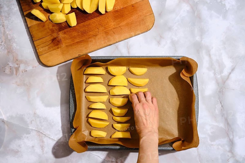 A hand is placing sliced potatoes on a baking tray lined with parchment paper. A wooden cutting board holds additional potato slices. This scene takes place in a kitchen with a clean work surface