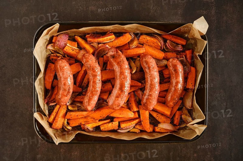 Natural casing sausages in a baking tray with sweet potatoes and onions. The tray contains roasted vegetables. The scene shows food preparation in a kitchen for a meal