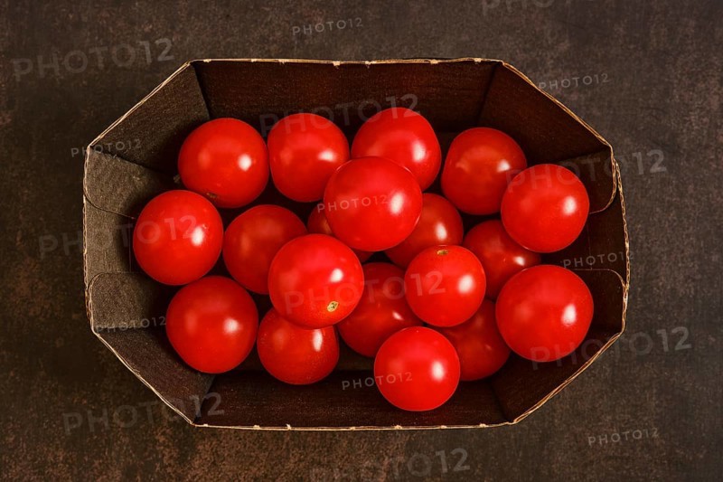 Cardboard box holds many red cherry tomatoes. The tomatoes are bright and round, placed together without any leaves or stems. The background is dark, making the colors stand out