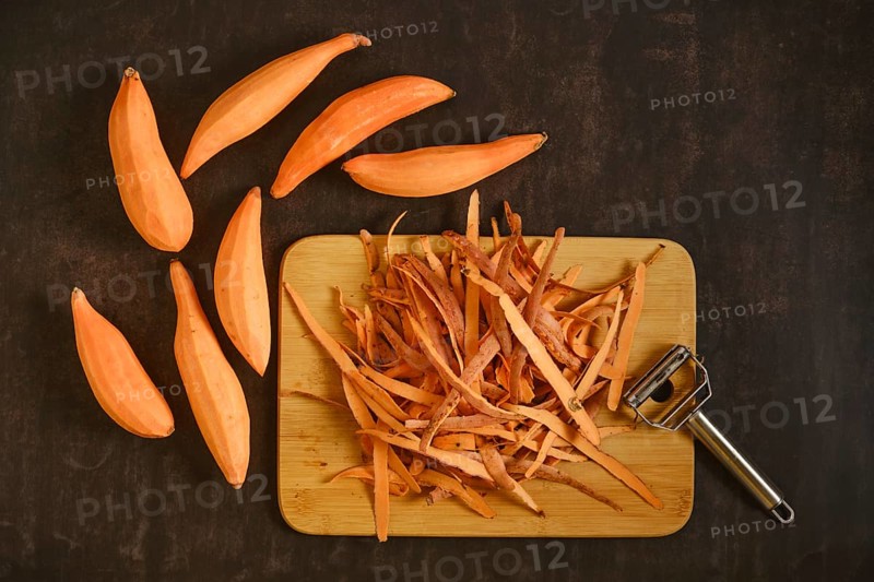 Peeled sweet potatoes with a vegetable peeler on a wooden cutting board. The skin is piled next to several whole sweet potatoes. The kitchen is tidy and organized