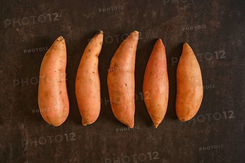 Five sweet potatoes are placed in a straight line on a dark surface. They vary slightly in size and shape. The setting is simple, emphasizing the natural appearance of the sweet potatoes