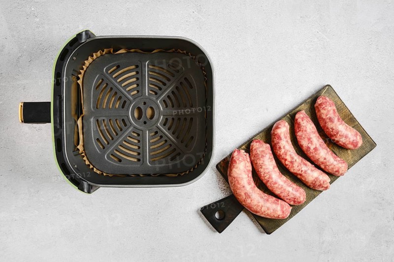 Fresh natural casing sausages on a cutting board beside an air fryer basket. The sausages are ready to be cooked for a meal. Preparation takes place in a kitchen setting