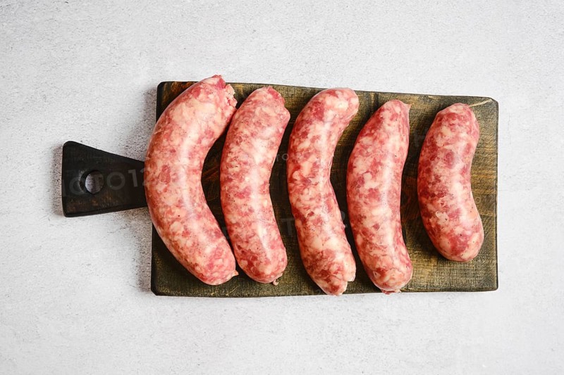 Five natural casing sausages on a wooden cutting board. They appear fresh and raw with visible meat and fat textures. These sausages are prepared for cooking or grilling