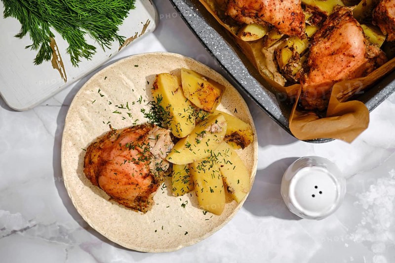 A plate holds pieces of chicken next to sliced and cooked potatoes. Fresh herbs are sprinkled on top. A tray with more chicken and potatoes is nearby. A salt shaker rests on the table