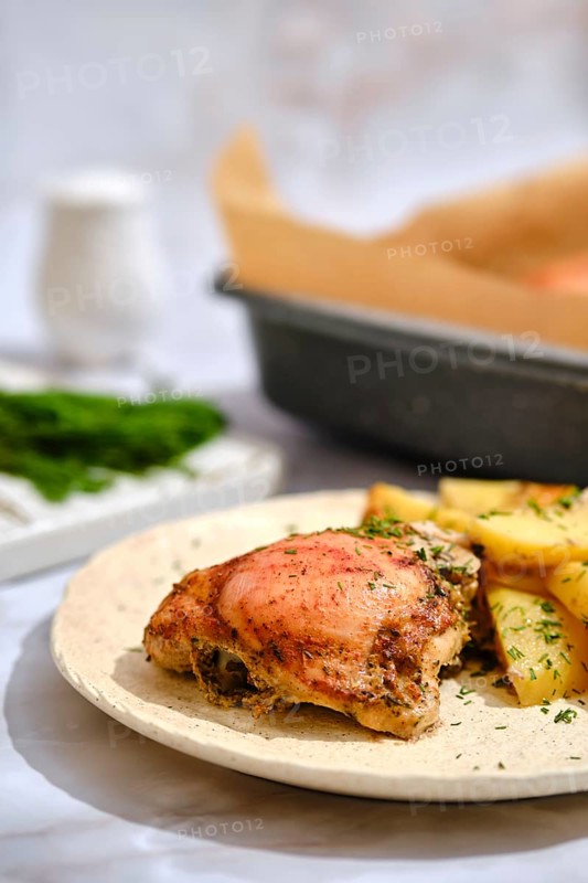 A plate holds a crispy chicken thigh next to golden roasted potatoes. Fresh green herbs are sprinkled on the food