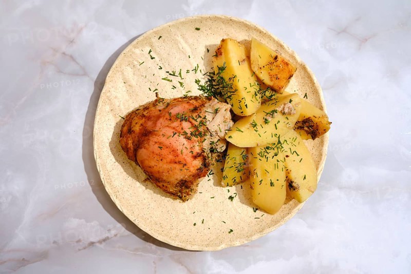 A chicken thigh is placed beside several pieces of seasoned potatoes on a round plate. The dish has a sprinkle of herbs on top. The setting is simple and focuses on the food