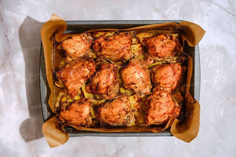Baking tray with seasoned chicken thighs and sliced potatoes. The dish was roasted in an oven, making a hearty meal for a family gathering. Ingredients are arranged neatly on a kitchen counter