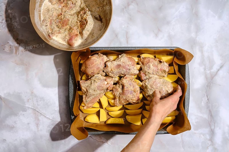 A person prepares a dish by placing seasoned chicken on a tray lined with potatoes. There is a bowl with marinade nearby. The setting is a kitchen, and the focus is on preparing a meal