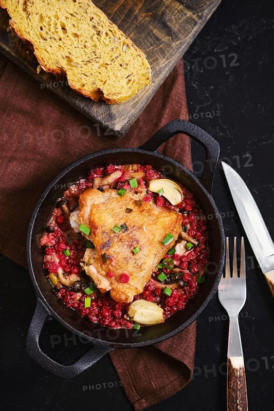 A warm chicken dish is served in a black cast iron pot. The chicken is on a bed of grains with herbs. Bread is placed on a wooden board nearby. Fork and knife rest beside the dish