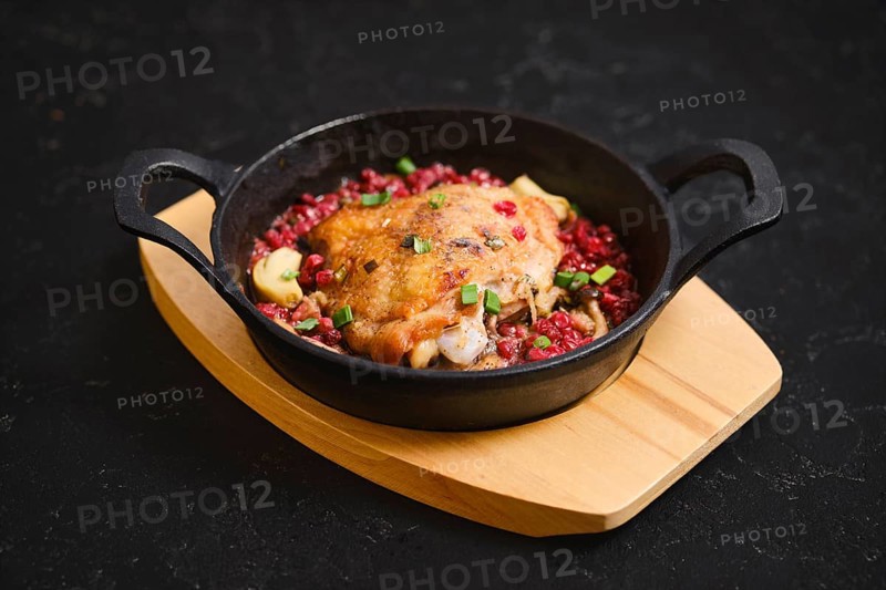 A cast iron skillet holds a serving of cooked chicken topped with lingonberry and chopped spring onion. The dish is placed on a wooden board. The setting is dark, creating focus on the food