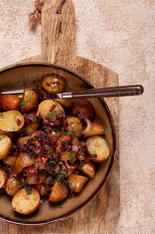 Succulent golden-brown roasted potatoes are served with sweet caramelized onions, garnished with fresh herbs, all beautifully arranged on a rustic wooden board in a warm kitchen
