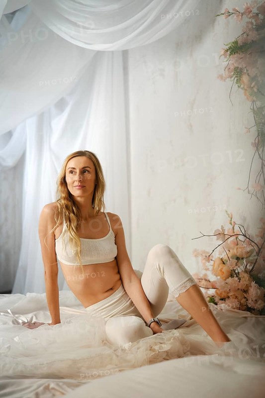 A woman in top and leggings sits on a bed and looks to her left. Soft light falls on her from above