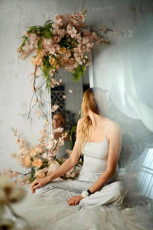 A woman sits cross-legged in a bright room. She faces a mirror decorated with flowers. Light filters through the window, creating a soft atmosphere. Her hair is long and flowing, adding to the scene