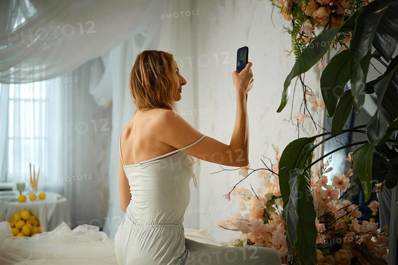 A woman sits in a room with light curtains and floral decorations, holding her phone to take a selfie. There are fruits displayed on a nearby table, adding color to the space