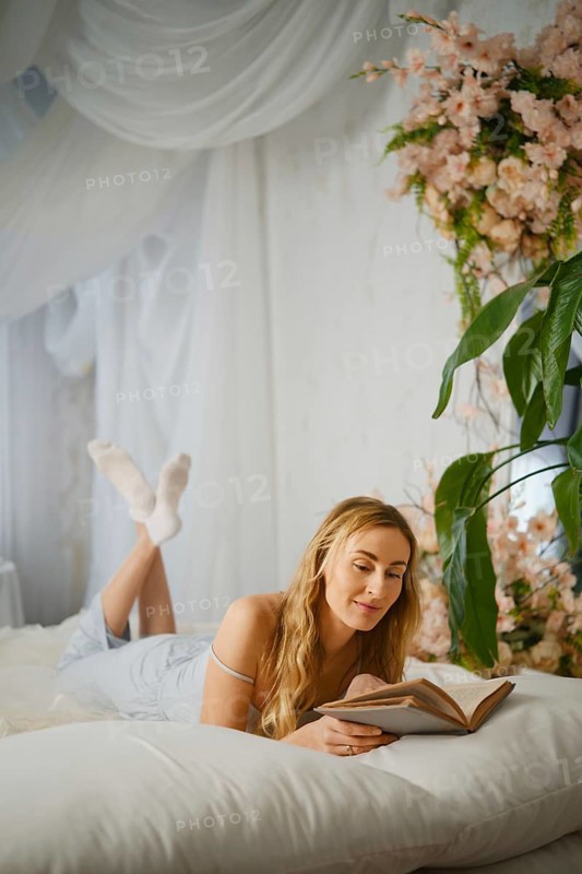 Reading a book while lying on a bed in a room with flowers and a plant during afternoon light