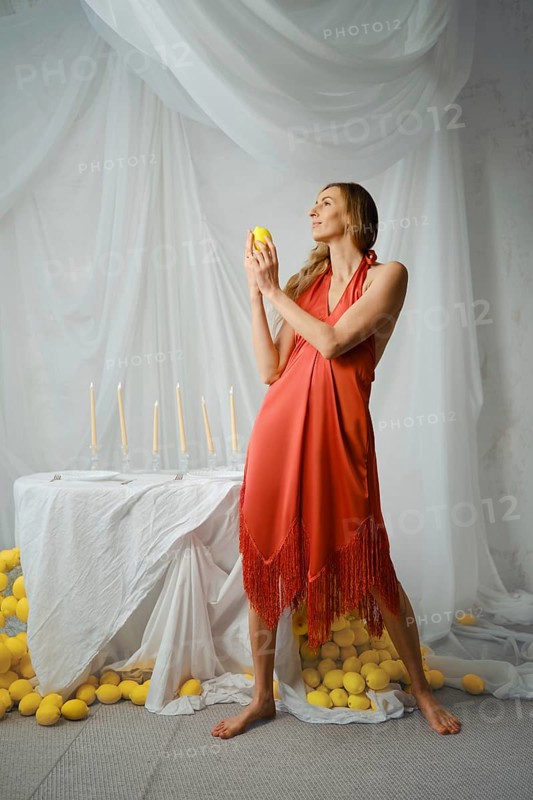 Woman stands in a studio holding a lemon while facing a table decorated with candles and surrounded by lemons. The background features draped fabric and the setting is bright and airy