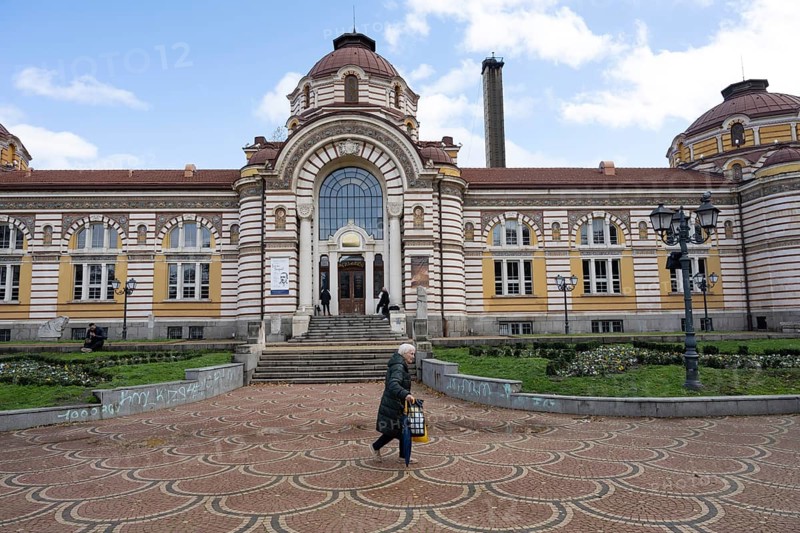 Sofia, Bulgaria. November 23rd 2025. An old woman passes the Regional History Museum of Sofia, the former Municipal Central Mineral Baths a historical landmark of Sofia, Bulgaria