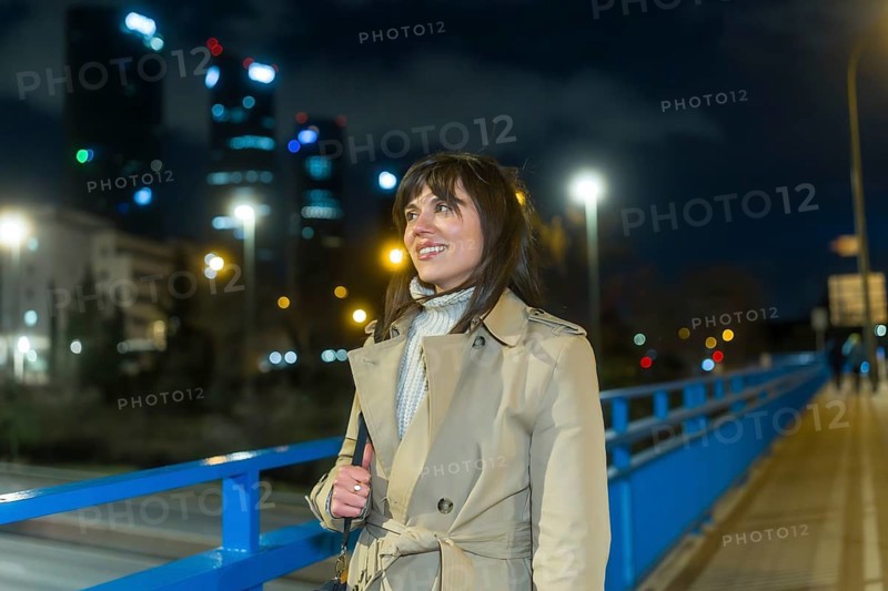 Woman wearing a trench coat and warm sweater standing on a bridge, looking at the illuminated city skyline with modern skyscrapers and night lights in an urban setting