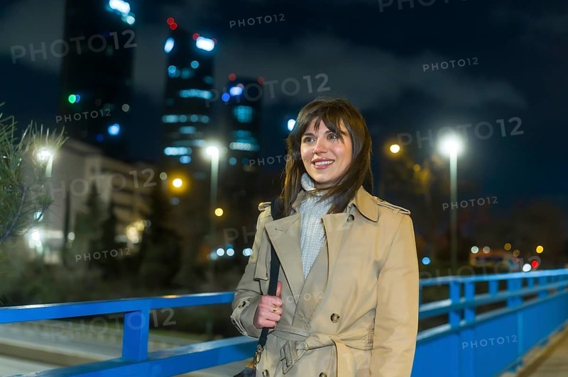 Young woman in a trench coat smiling as she walks across a city bridge at night, skyline lights and illuminated skyscrapers creating a warm bokeh urban backdrop