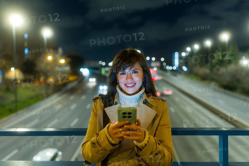 Young woman standing on an urban bridge at night, smiling while reading messages on her mobile phone, with blurred car lights and city streets creating a dynamic backdrop