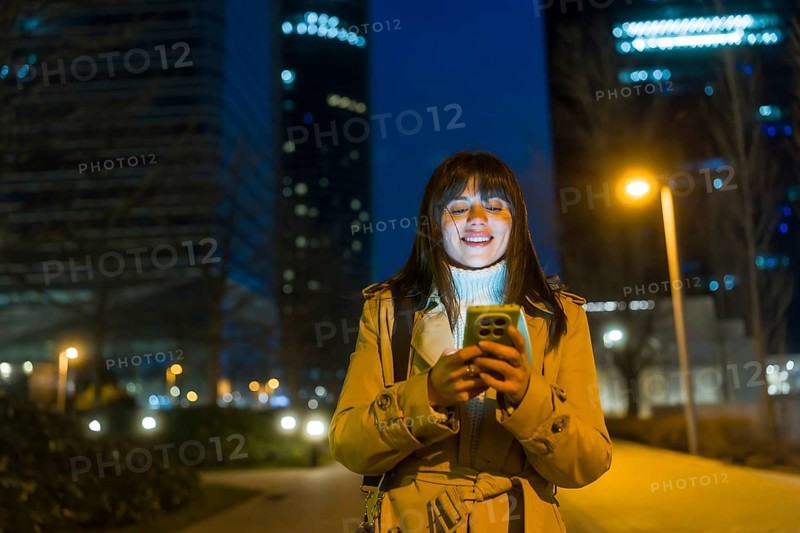 Young woman standing on an urban street at night, looking at her smartphone and smiling, with technology illuminating her face against a backdrop of blurred city lights and buildings