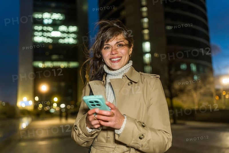 Young woman in a trench coat stands on a lit city street at dusk, smiling at the camera while holding a smartphone, conveying modern connectivity and urban lifestyle