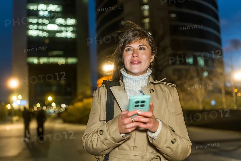 Woman standing outdoors at night, using a smartphone for communication and networking, dressed in a trench coat with city lights and blurred buildings creating an urban background