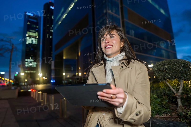 Woman stands at twilight in a glowing downtown, smiling while using a tablet, blurred skyscrapers and lit streets suggest urban tech lifestyle, business and connectivity