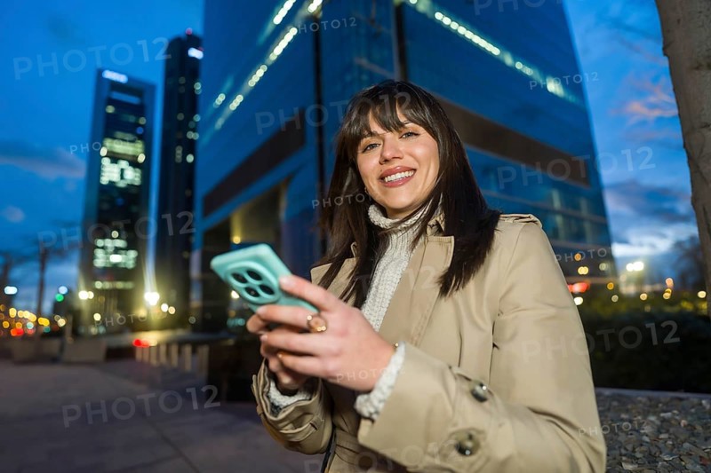Young professional woman smiling while using her smartphone for networking and social media in a modern urban business district at dusk, confident and connected outdoors