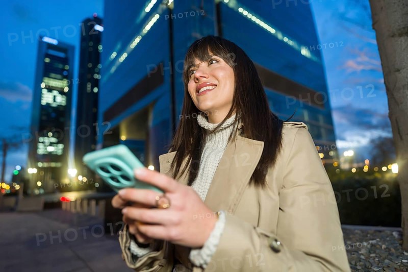 Young professional woman smiling and looking away while interacting with a smartphone in a modern city at night, reflecting technology, connectivity, and urban living