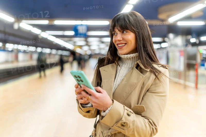 Woman standing on a train station platform, smiling and looking away while sending messages or browsing on her smartphone, representing urban connectivity and modern commuting
