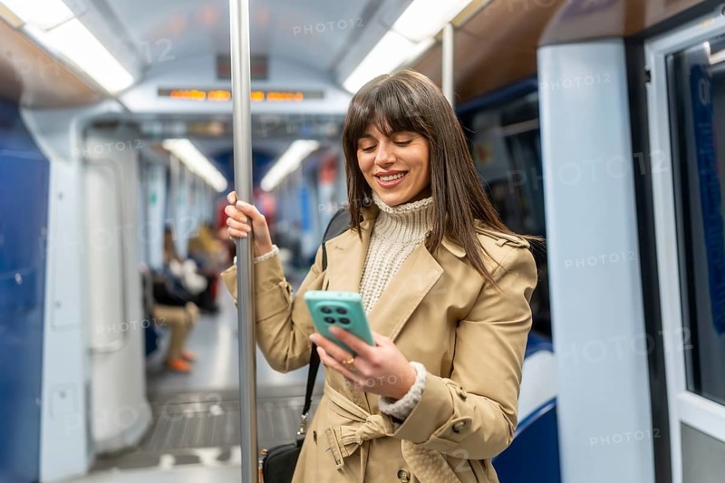 Young woman standing in a subway car, smiling at her smartphone screen during the commute, browsing messages and social media while holding the pole on a busy urban train