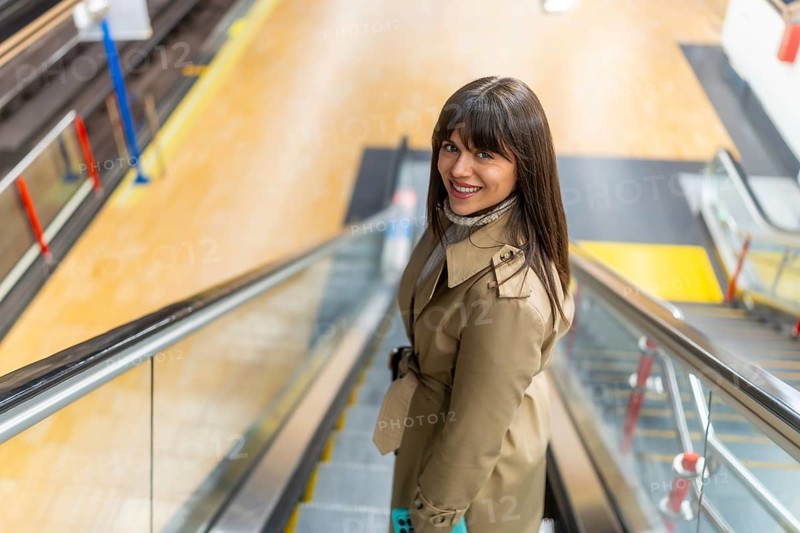 Woman in a trench coat smiling confidently while riding down an escalator in a modern subway station, representing urban lifestyle, daily commute, and public transportation