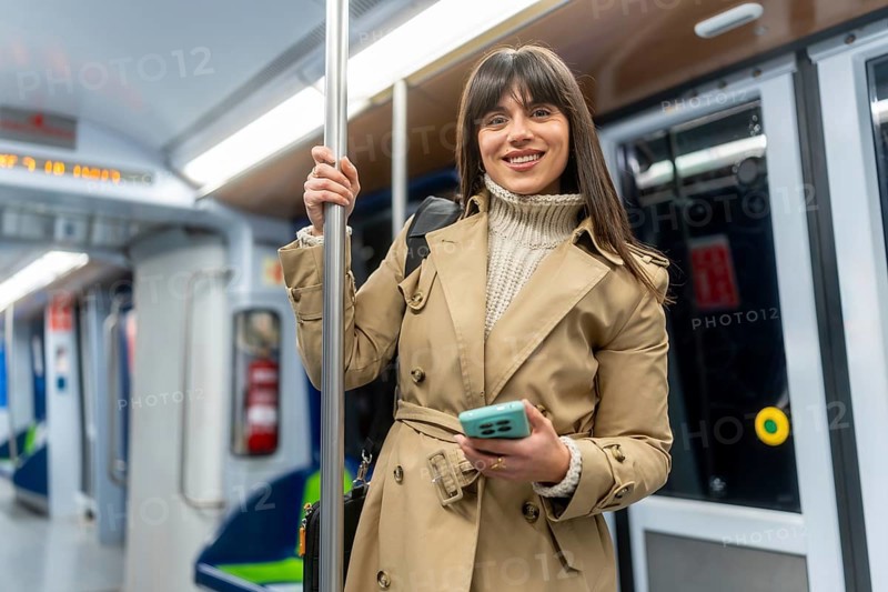 Young smiling woman in a trench coat stands in a subway car, holding her smartphone while commuting through the city, scrolling and enjoying her urban journey