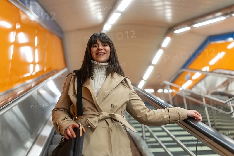 Woman in a trench coat and sweater ascending an escalator in a modern subway station, holding a bag and looking at the camera with a cheerful expression, symbolizing urban commute and daily travel
