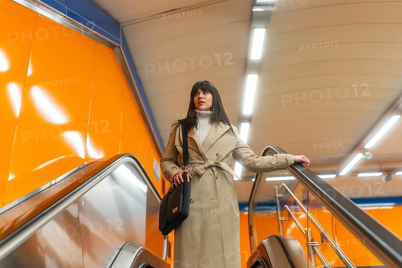 Woman travelling on an escalator in a subway station, holding onto the handrail, dressed in a trench coat, reflecting urban movement, daily routine, and public transportation