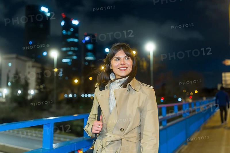 Woman in trench coat on an urban bridge at night, smiling and looking away as illuminated skyscrapers and city lights of madrids cuatro torres sparkle in the evening haze