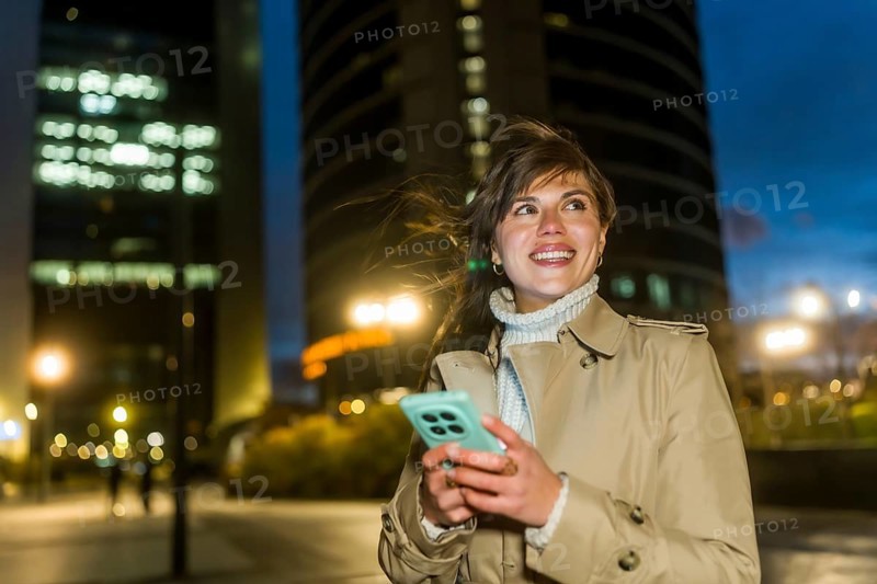 Young woman smiling, looking up while navigating her smartphone, dressed in a trench coat and turtleneck, enjoying a pleasant stroll through a modern city district during the evening