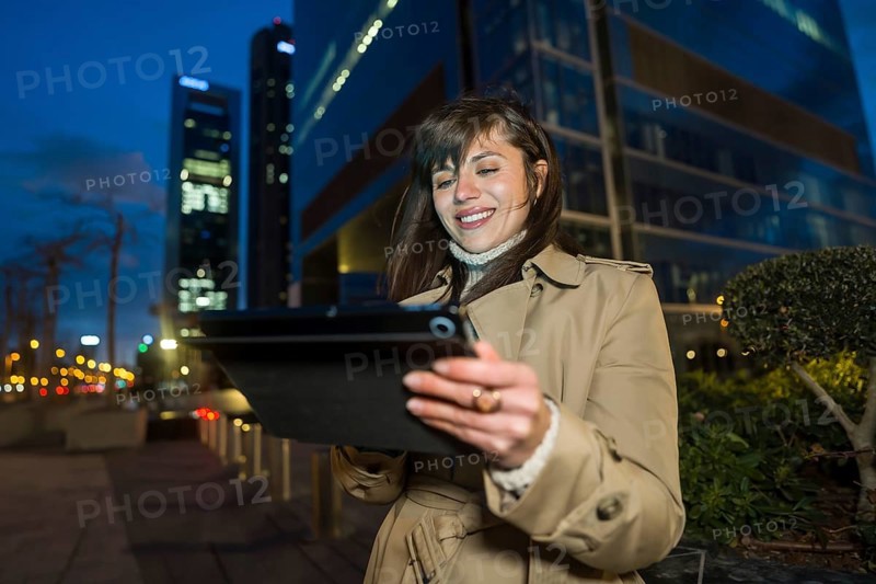 Smiling young businesswoman wearing a trench coat standing outdoors at night, connecting with a digital tablet in front of modern office buildings in the urban illuminated cityscape