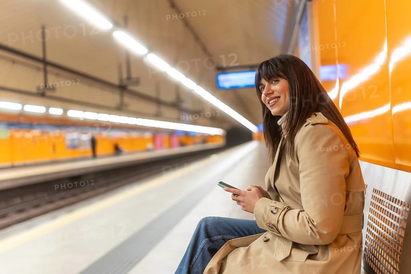 Young woman commuting, checking her mobile phone and smiling while sitting on a bench at an empty underground subway platform, enjoying her transit journey