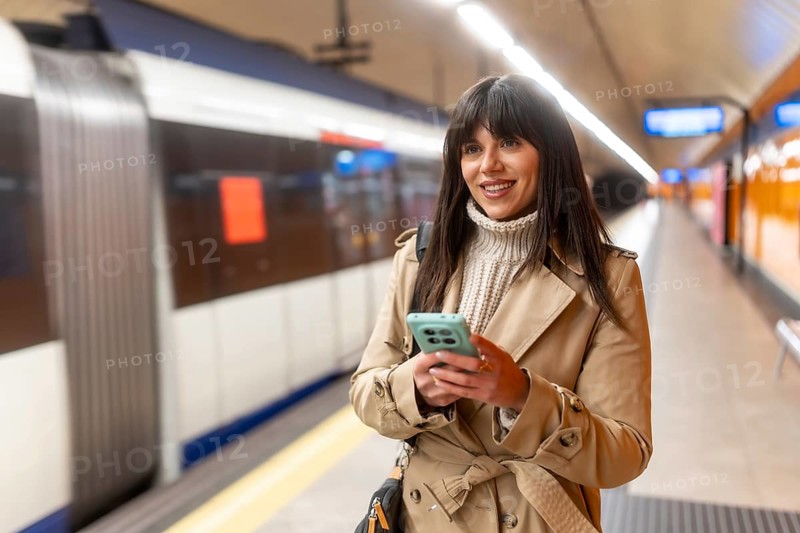 Young woman standing on a subway platform, wearing a trench coat and sweater, happily interacting with her mobile phone as she waits for public transport