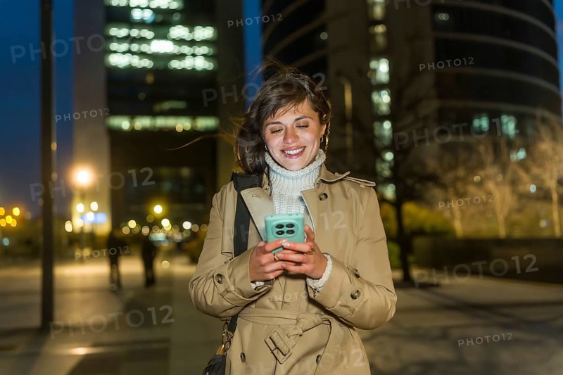 Young woman in a trench coat smiling, walking through a city at night, connecting with her smartphone, experiencing urban lifestyle and happy communication under city lights